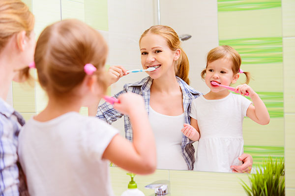 Young girl and her mom brushing teeth while looking in the mirror.