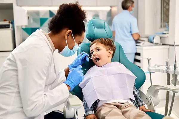 A young child sits calmly in the dental chair on his own, opening his mouth for his pediatric dentist to perform a routine dental examination using common dental tools.