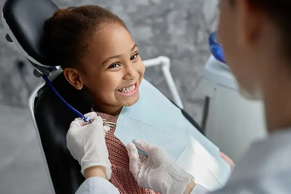 A smiling, excited young girl sits in the dental chair, eagerly awaiting her dental exam while her dentist places a bib on her.