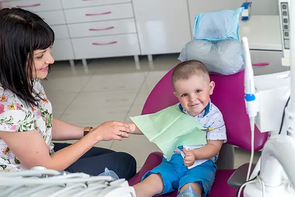 A little boy wearing a bib comfortably waiting for the dentist with his mom nearby.
